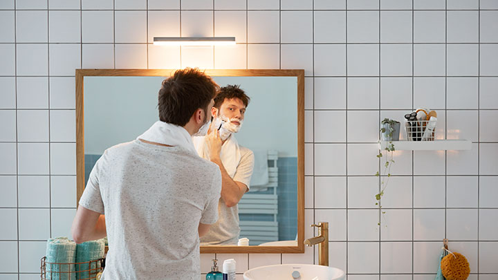 Un homme se rase devant un miroir de salle de bains éclairé par une ampoule à DEL Philips fixée au mur