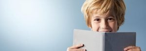 "Young child with blonde hair smiling behind open book, showcasing joyful reading experience against light blue background."