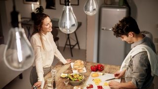 Une femme observe un homme couper des légumes sur une planche à découper dans une cuisine, vous pouvez voir des ampoules dans la partie supérieure de l’image
