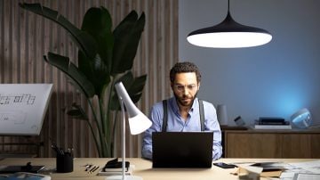 Un homme est assis à un bureau avec un portable devant lui, éclairé par un luminaire suspendu au-dessus et une lampe de table à sa droite